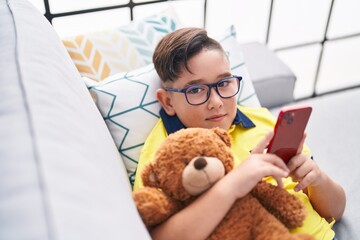 Sticker - Adorable hispanic boy using smartphone hugging teddy bear at home