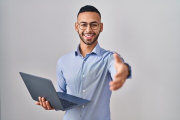 Poster - Young hispanic man working using computer laptop smiling friendly offering handshake as greeting and welcoming. successful business.