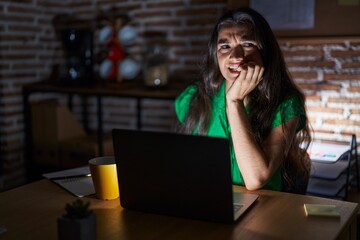 Young teenager girl working at the office at night looking stressed and nervous with hands on mouth biting nails. anxiety problem.