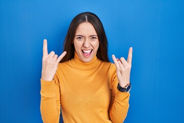 Poster - Young brunette woman standing over blue background shouting with crazy expression doing rock symbol with hands up. music star. heavy music concept.