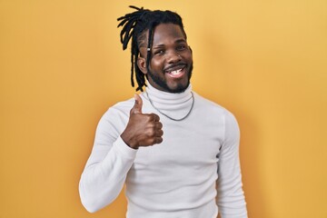 Sticker - African man with dreadlocks wearing turtleneck sweater over yellow background doing happy thumbs up gesture with hand. approving expression looking at the camera showing success.