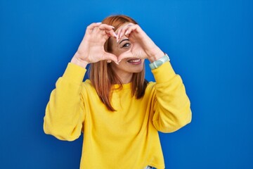 Wall Mural - Young woman standing over blue background doing heart shape with hand and fingers smiling looking through sign