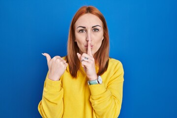 Wall Mural - Young woman standing over blue background asking to be quiet with finger on lips pointing with hand to the side. silence and secret concept.