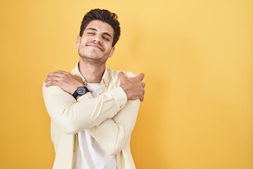 Sticker - Young hispanic man standing over yellow background hugging oneself happy and positive, smiling confident. self love and self care