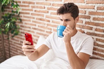 Wall Mural - Young hispanic man using smartphone drinking coffee at bedroom