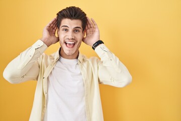 Poster - Young hispanic man standing over yellow background smiling cheerful playing peek a boo with hands showing face. surprised and exited