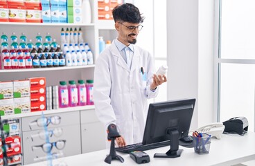 Poster - Young hispanic man pharmacist holding pills bottle using computer at pharmacy