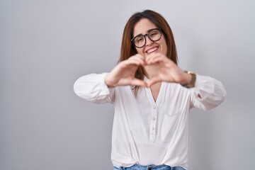 Wall Mural - Brunette woman standing over white isolated background smiling in love doing heart symbol shape with hands. romantic concept.