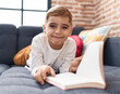 Adorable hispanic boy reading book lying on sofa at home