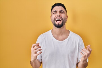 Sticker - Young handsome man wearing casual t shirt over yellow background crazy and mad shouting and yelling with aggressive expression and arms raised. frustration concept.