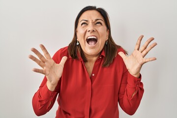 Sticker - Hispanic mature woman standing over white background crazy and mad shouting and yelling with aggressive expression and arms raised. frustration concept.