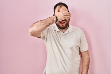 Wall Mural - Plus size hispanic man with beard standing over pink background smiling and laughing with hand on face covering eyes for surprise. blind concept.