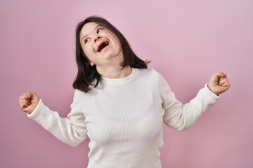 Poster - Woman with down syndrome standing over pink background celebrating surprised and amazed for success with arms raised and eyes closed. winner concept.