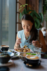 Poster - A young Asian woman sits eating pasta at a table in a cafe.