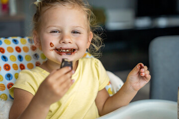 Wall Mural - Little girl with blond hair eating homemade chocolate with dirty mouth and hands in home kitchen