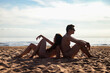 Silhouettes of naked couple man and woman sitting back to back on sandy nudist sea beach outdoors. Nude young naturist couple relaxing, idyllic. Nudism naturism lifestyle concept. Copy ad text space