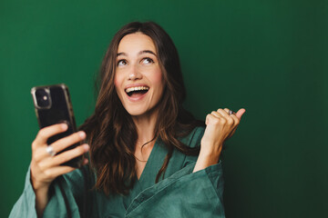 Fun young woman hold in hand use mobile cell phone doing winner gesture clenching fists isolated on green background studio. Girl using smartphone apps winning online celebrating discount gift voucher