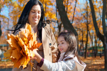 Poster - mother and daughter are in autumn city park, happy people walking together, family with children, playing with yellow leaves, beautiful nature, bright sunny day