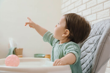 Cute little baby pointing at something in high chair indoors