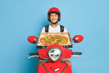 Wall Mural - Delivery boy wearing red helmet posing inside studio on his red scooter and holding pizza box in hands, fast delivery concept, copy space