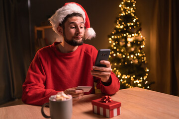 Dissatisfied man to make a purchase in an online internet store, man surprised on Christmas, sitting at the table with smartphone and credit card at home near Christmas tree