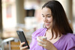 © PheelingsMedia - Happy female checking phone in a coffee shop terrace