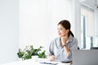 Charming asian businesswoman sitting looking away out of window hand holding pen working on laptop in office. copy space