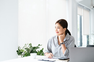 Poster - Charming asian businesswoman sitting looking away out of window hand holding pen working on laptop in office. copy space