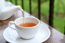 Pouring Hot Tea From White Ceramic Teapot Into Porcelain Cup. Outdoor Breakfast On Balcony Cafe.