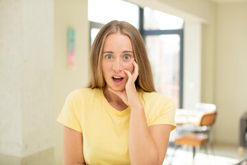 Wall Mural - pretty blond woman open-mouthed in shock and disbelief, with hand on cheek and arm crossed, feeling stupefied and amazed