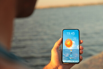 Wall Mural - Man checking weather using app on smartphone near river, closeup. Data, sun and other illustrations on screen