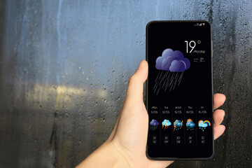 Wall Mural - woman checking weather using app on smartphone near wet window, closeup. data, cloud with rain and o