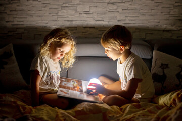 Poster - Two blond cute childrem, boy and girl, siblings, lying under the cover in bed, reading book together with small light