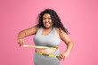 © Prostock-studio - Smiling black overweight woman in sportswear checking waist with measuring tape, isolated on pink studio background