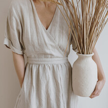 Young Beautiful Woman In Neutral Beige Creamy Linen Dress Holding Sandy Earthenware With Dried Pampas Grass Over White Wall