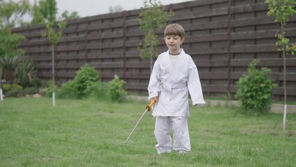 Wall Mural - Funny little boy barefoot in white karate suit plays with sword in the garden