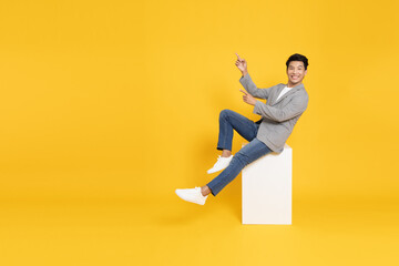 Wall Mural - Young Asian businessman sitting and pointing to empty copy space isolated on yellow background