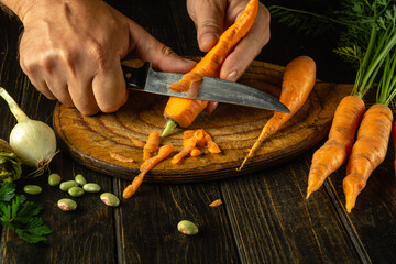 Wall Mural - The cook cleans fresh carrots on the kitchen table before adding them to vegetable borscht. Close-up of chef hands with knife while peeling carrot
