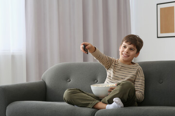 Poster - Little boy holding bowl of popcorn and changing TV channels with remote control on sofa at home