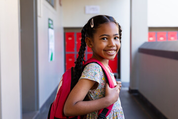 Portrait of happy biracial schoolgirl with school bag smiling in corridor at elementary school