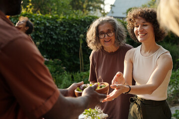 Focus on young cheerful brunette woman taking bowl with food from hands of her husband helping her with serving table for family dinner