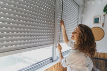 Young Woman Opening Roller Blind Shutters On The Balcony On A Summer Day