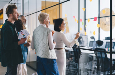 Wall Mural - Group of experienced office workers talking while collaborating on informative memo sticked to glass board in office interior, confident male and female partners briefing and communicating in workshop