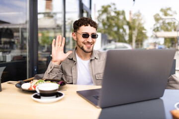 Wall Mural - Handsome man waving hand to laptop web camera while chatting with family online at street cafe. Video call concept