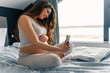 © DusanJelicic - A pregnant woman with long brown hair is sitting on the bed in front of the window with her legs crossed with her hand gently on her stomach while using her phone to take pictures