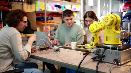 young happy engineers programming an yellow robotic arm in the workshop to grab cardboard water glas