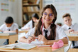 © AnnaStills - Portrait of schoolgirl in eyeglasses smiling at camera while sitting at her desk at lesson