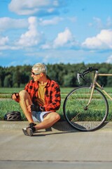 a guy in glasses and a red shirt is resting on the side of the road taking off his bicycle helmet, a vintage sports bike is standing nearby