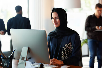 Selective focus on Emirati National woman in Abaya and Hijab at office using desktop computer display screen with blurred workmates as background. Emiratisation concept in the United Arab Emirates