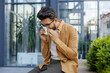 © Liubomir - Young man sneezing and having a runny nose allergy sitting on a bench in the daytime outside an office building, hispanic businessman sick with a tissue near his nose.